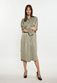 Olivengrünes Satinhemdkleid mit verdeckter Knopfleiste, Brusttasche und langen Ärmeln, kombiniert mit schwarzen High Heels.