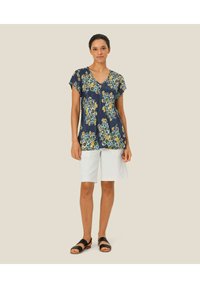 Marineblå top med blomsterprint i blå og gule nuancer, med korte ærmer og v-halsudskæring, parret med hvide shorts og sorte sandaler.