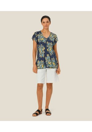Marineblå top med blomsterprint i blå og gule nuancer, med korte ærmer og v-halsudskæring, parret med hvide shorts og sorte sandaler.