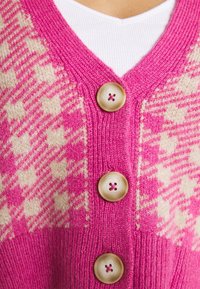 Růžový pletený cardigan se vzorem houndstooth v krémové barvě, třemi velkými krémovými knoflíky a výstřihem do V. Měkká textura.