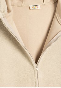 Hoodie zippé en polaire beige avec un extérieur texturé, un intérieur uni et une étiquette indiquant la taille et la marque, convenant aux enfants de 9 à 10 ans.