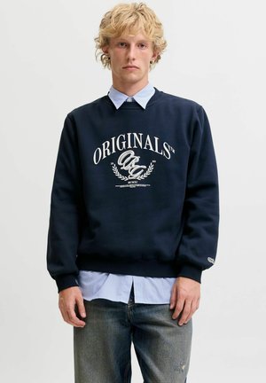 Jeune homme aux cheveux blonds et bouclés portant un sweat bleu marine portant l’étiquette "Originals" par-dessus une chemise bleue à col et un jean délavé.