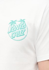 Μπροστινή πλευρά λευκού μπλουζακιού με τυρκουάζ λογότυπο "Santa Cruz Skateboards" που παρουσιάζει δύο φοίνικες μέσα σε κύκλο.