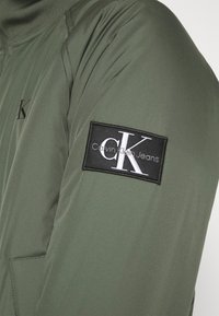 Groene jas met een gladde textuur, voorzien van een zwarte patch op de mouw met de tekst "Calvin Klein Jeans" en een CK-logo.