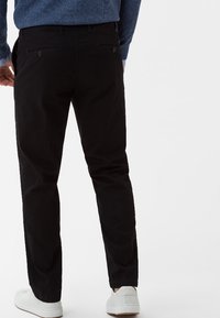Muž oblečený v černých slim-fit kalhotách, modrém svetru a bílých teniskách, stojící zády k fotoaparátu na bílém pozadí.