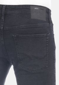 Vaqueros de mezclilla negra con una textura suave, presentando un parche de cuero en la parte trasera, corte recto y bolsillos traseros estándar con un sutil detalle de costura.