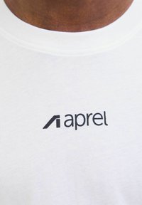 Balts kokvilnas T-krekls ar melnu logotipu, uz kura ir rakstīts "aprel," centrāli izvietots krūšu daļā. Vienkāršs, gluds dizains.