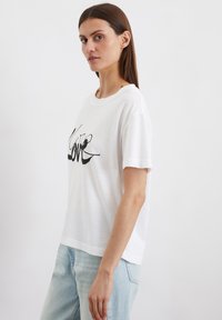 Weißes Baumwoll-T-Shirt mit rundem Halsausschnitt und kurzen Ärmeln. Mit einem schwarzen Grafikdruck, der "Love" über die Vorderseite sagt.