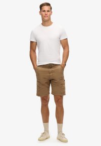 Brune cargo shorts med flere lommer, parret med en tætsiddende hvid t-shirt. Skoene er beige med lysegrå sokker. Ingen bemærkelsesværdige mønstre.