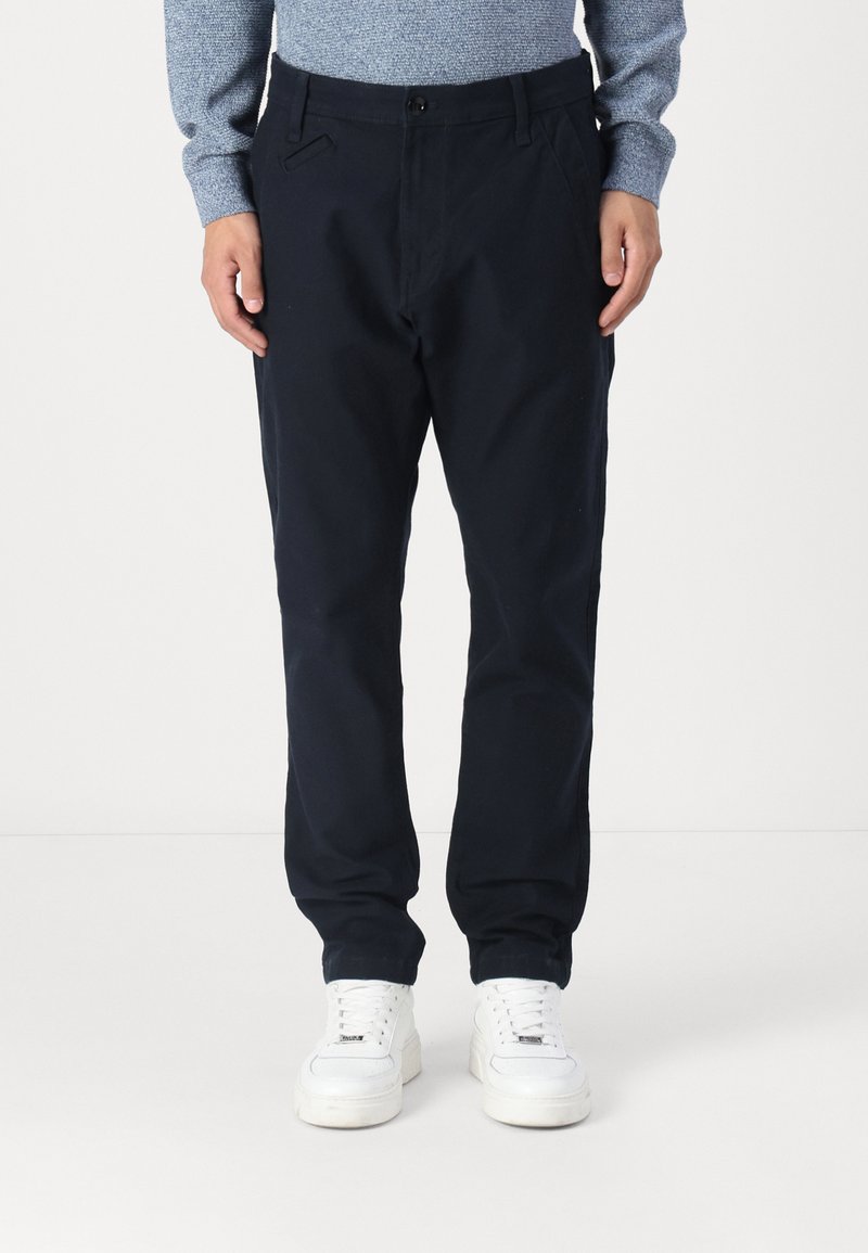 Navy blue chinos made of cotton blend fabric, featuring a straight-leg design, side pockets, and a button closure, paired with white sneakers.