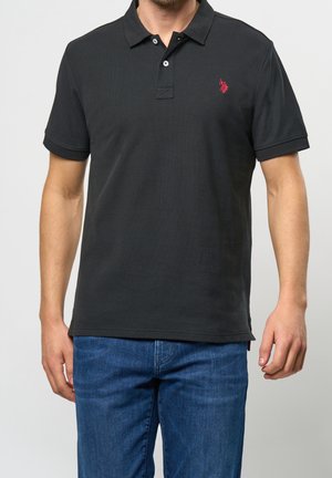 Polo noir en tissu texturé, doté d'une patte de boutonnage à deux boutons et d'un petit logo rouge sur la poitrine gauche. Associé à un jean en denim bleu.