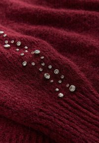 Burgundy stickad tröja med en texturerad yta där en rad av klara strassmarker formar ett dekorativt mönster längs ena sidan.