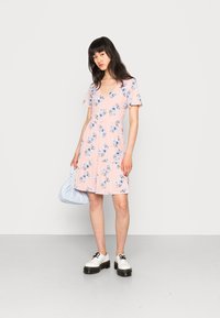 Vestido de flores em rosa claro, decote em V, mangas curtas, saia em camadas, feito de um tecido suave. Combinado com sapatos brancos robustos e uma bolsa azul clara.