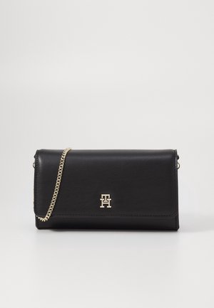 Sort læderclutch med guldkæde og guld "T" logo på frontklappen, sat mod en ensartet lys baggrund.