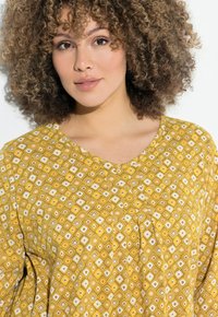Blouse jaune à motifs avec un design géométrique, des textures douces, un décolleté en V et des détails froncés au niveau de la poitrine.