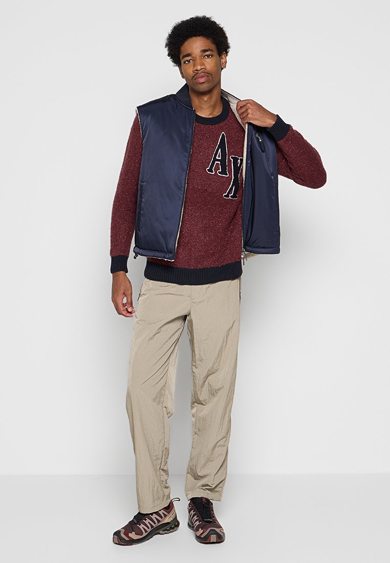 Gilet smanicato blu navy sopra un maglione lavorato a maglia bordeaux con logo "AX", abbinato a pantaloni beige chiaro e sneakers camouflage.