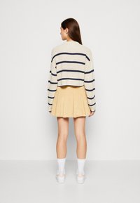Sweater blanc à côtes avec des rayures bleu marine, associé à une jupe plissée jaune. Le modèle porte des chaussettes blanches et des chaussures, de dos face à l'appareil photo.
