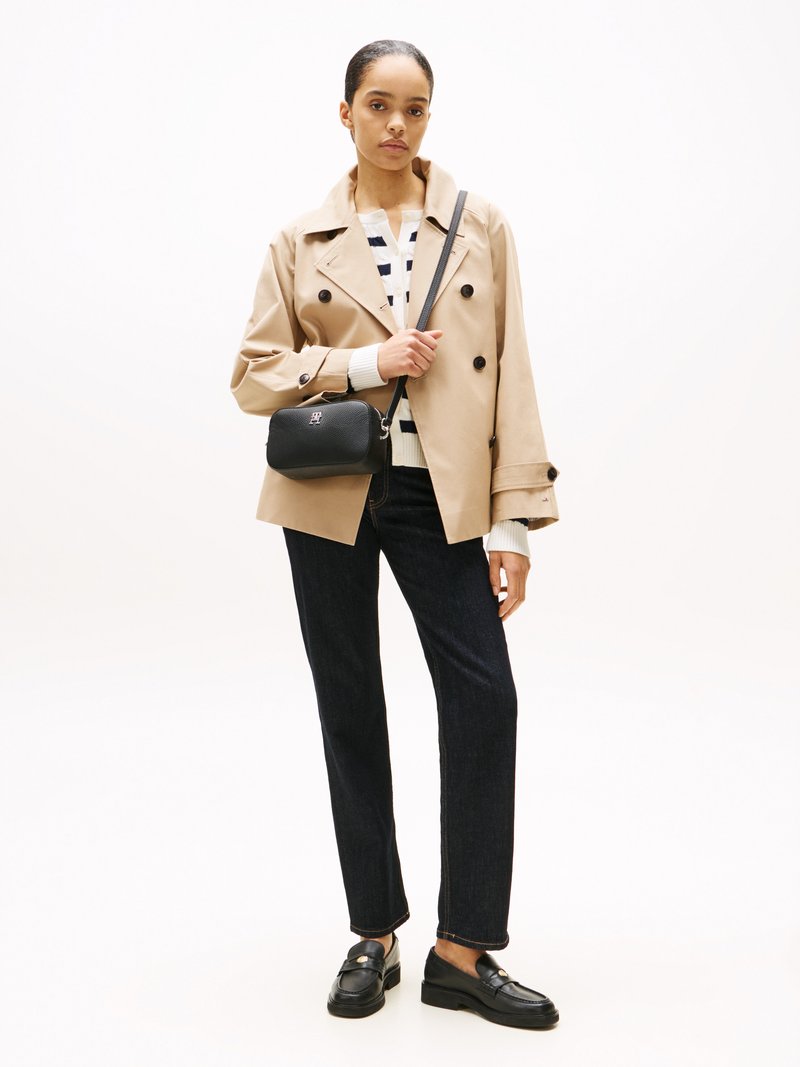 Donna che indossa un cappotto doppio petto beige, un maglione a righe, jeans scuri, mocassini neri e una borsa a tracolla nera, in piedi contro uno sfondo chiaro.