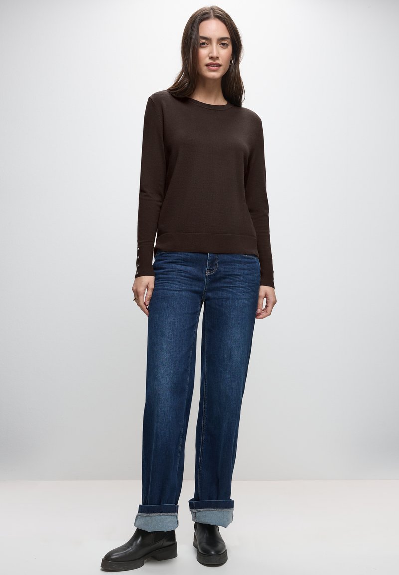 Frau steht vor einem einfarbigen Hintergrund und trägt einen braunen Pullover mit langen Ärmeln, hochgekrempelte blaue Jeans und schwarze Stiefeletten.