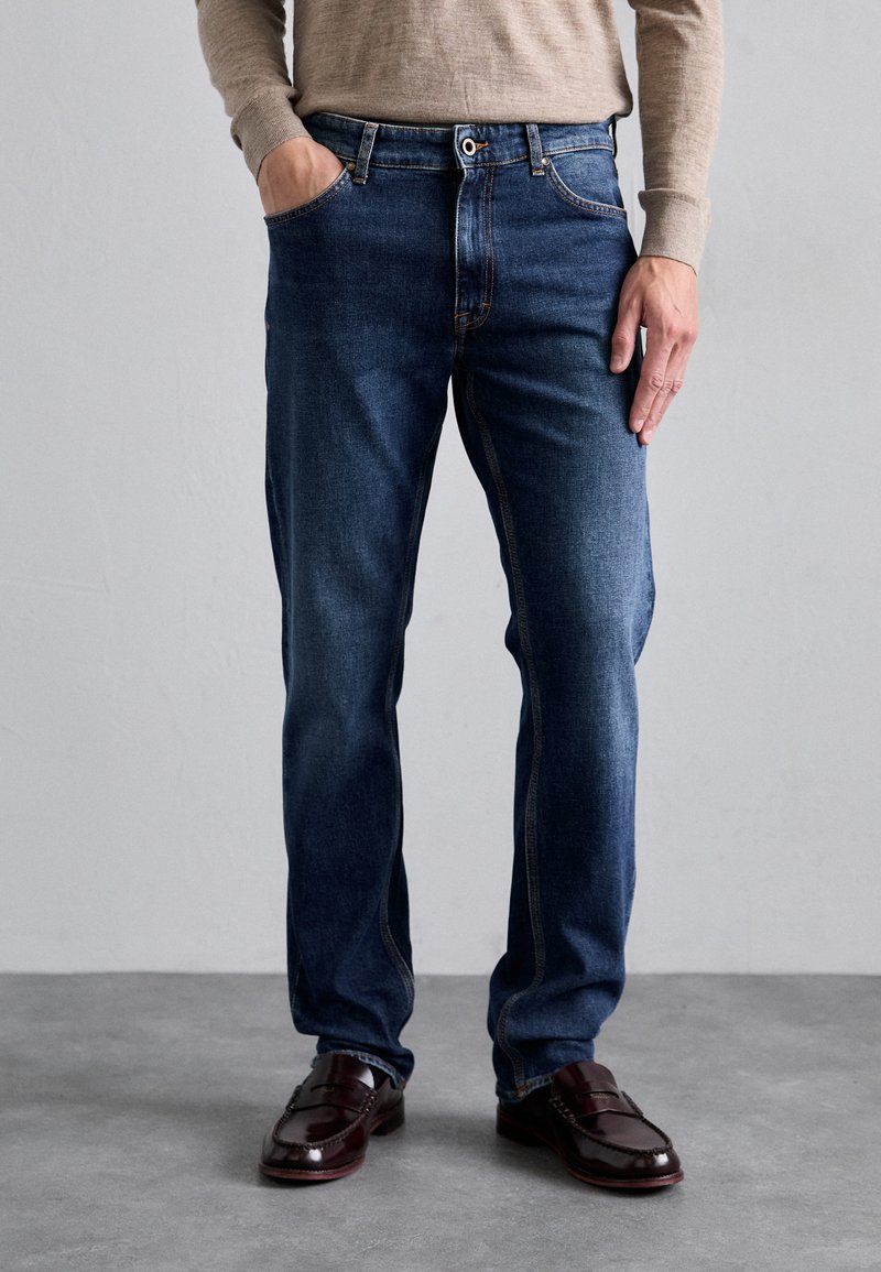 Jean en denim bleu foncé avec une coupe droite, présentant un design classique à cinq poches et des coutures orange contrastantes. Porté avec des chaussures en cuir foncé.