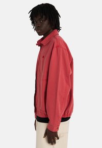 Młoda osoba z ciemnymi dredami, ubrana w czerwony dresowy sweter na zamek i beżowe spodnie, stojąca bokiem na jednolitym tle.