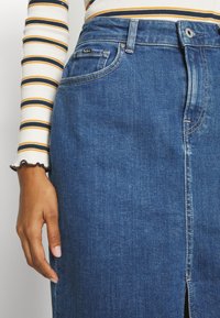 Hand som vilar bredvid en högmidjad blå denimkjol med en frontslits, parat med en vit långärmad tröja med senapsgula och marinblå ränder.