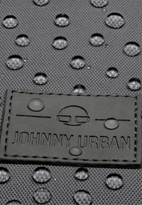 Mustat vedenkestävät kankaat, joissa on kohotettu rakenne, varustettuna kohokuvioidulla "JOHNNY URBAN" -merkinnällä ja vesipisaroilla pinnalla.