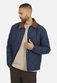 Marine gewatteerde jas met een bruine corduroy kraag, drukknoopsluitingen en elastische manchetten. Draag over een lichtbeige overhemd.