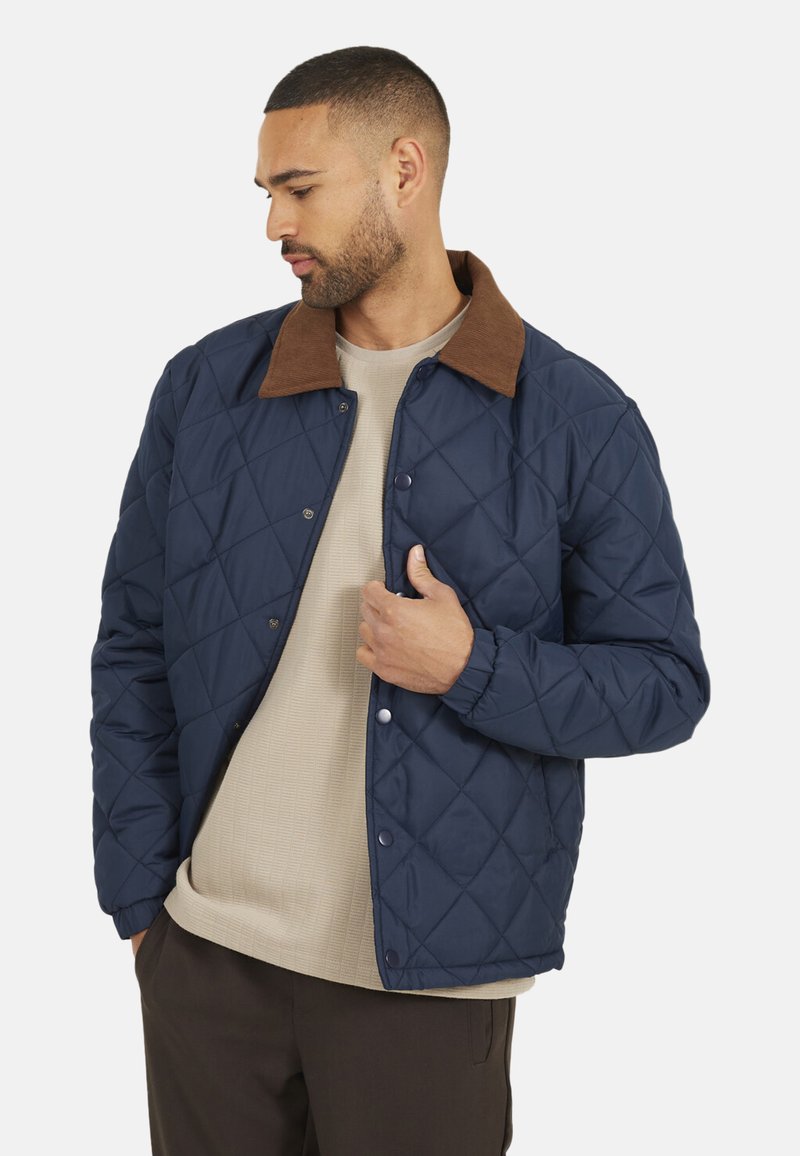 Marine gewatteerde jas met een bruine corduroy kraag, drukknoopsluitingen en elastische manchetten. Draag over een lichtbeige overhemd.