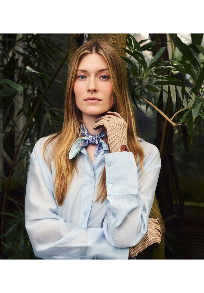 Mujer rubia con camisa azul abotonada y pañuelo estampado en el cuello, de pie con los brazos cruzados, usando guantes de punto beige, frente a un fondo de hojas verdes.
