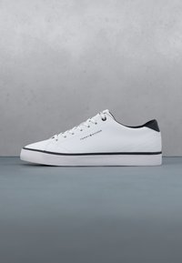 Baskets basses blanches avec talon noir, bordure noire, lacets blancs et logo Tommy Hilfiger sur le côté sur fond gris.