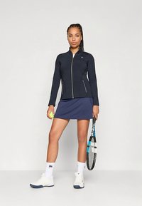 Mörkblå idrottsjacka med dragkedjefickor, i kombination med en matchande mörkblå kjol. Tillbehör inkluderar ett tennisracket och en grön tennisboll.