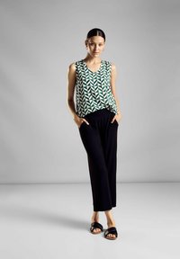 Top senza maniche con un motivo geometrico verde, abbinato a pantaloni larghi neri. Il modello sta in piedi, con le mani nelle tasche, indossando sandali neri.