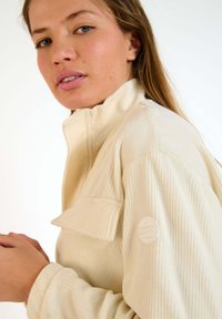 Pullover in tessuto a costine colore crema con colletto alto, dotato di tasca sul petto e superficie testurizzata. Logo del marchio sulla manica.