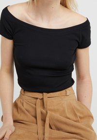 Svart, off-the-shoulder, kortärmad topp gjord av mjuk tyg, tillsammans med högmidjade, bruna, texturerade kjol med bälte.