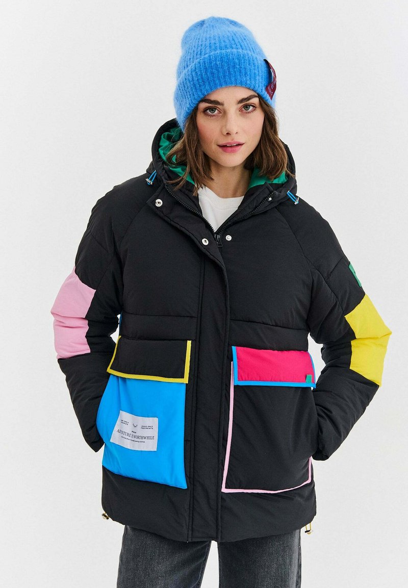 Žena v čiernej prešívanej bunde s farebnými vreckami a detailmi, s modrou pletenou čiapkou, stojaca na jednoliatom bielom pozadí.