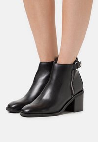 Botas de tobillo de cuero negro con una textura suave, un cierre de cremallera lateral y un tacón grueso. Presentan un diseño minimalista y una correa en el tobillo.