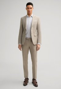 Completo beige su misura con una finitura strutturata, pantaloni coordinati e una camicia bianca. Scarpe eleganti marroni completano il look formale.