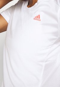 Vit atletisk t-shirt i andningsbart tyg, med korta ärmar, rund halsringning och en diskret meshpanel med en röd Adidas-logotyp.