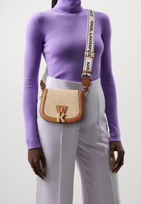 Henkilö, jolla on päällään violetti poolopaita ja vaaleanharmaat housut, kantaa vaaleanruskeaa kudottua olkalaukkua, jossa on "K"-riipus ja merkkihihna.