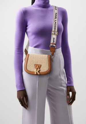 Henkilö, jolla on päällään violetti poolopaita ja vaaleanharmaat housut, kantaa vaaleanruskeaa kudottua olkalaukkua, jossa on "K"-riipus ja merkkihihna.