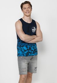 Mornarsko modra in modra tie-dye brezrokavnik s tiskom lobanje; v kombinaciji s sivo uničenimi kratkimi hlačami z raztrganinami in ravnim krojem. Standardna prilega.