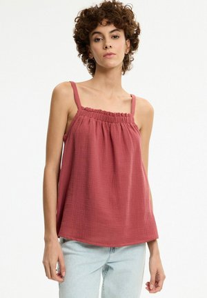 Mujer con cabello corto y rizado, que lleva una camiseta roja sin mangas con textura y vaqueros azules claros, de pie contra un fondo blanco liso.