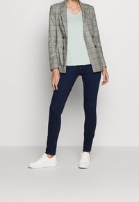 Blazer de cuadros gris de doble botonadura sobre una blusa verde claro con escote en V, combinado con jeans ajustados azul oscuro y zapatillas blancas.