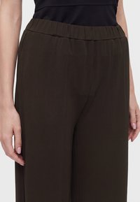 Taille et parties supérieures des jambes d'une personne portant un pantalon large taille élastique brun foncé et un haut noir, mains détendues sur les côtés.