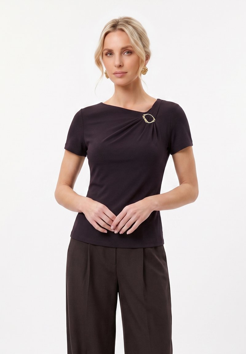 Femme blonde portant un haut noir à manches courtes avec une boucle décorative dorée près de l'encolure et un pantalon plissé foncé, debout immobile.
