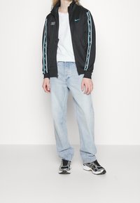 Svart atletisk jacka med turkosa detaljer, dragkedja framtill och Nike-logo, kombinerad med ljusblå jeans med raka ben och svarta sneakers.