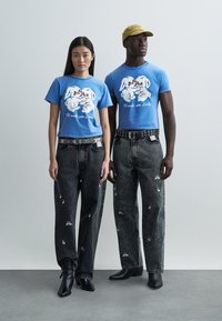 Två modeller står bredvid varandra. De bär matchande blå t-shirts med tecknade hundar. Ett par slitna svarta jeans med vita mönster.