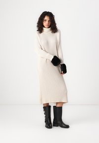 Cream ribbed sweater dress with high neckline, black fur accents on wrists, paired with knee-high black leather boots featuring buckles.