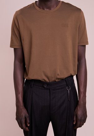 Mørk hudfarvet person iført en brun Hugo Boss t-shirt stoppet ned i sorte bukser med nålestriber og et tilbehør med nøgleringsløkke.